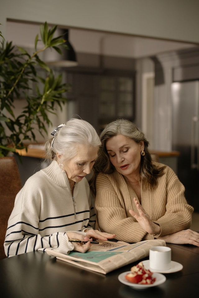 Two elderly women sitting at a table looking at a newspaper, seeking wellness tips for their senior health.