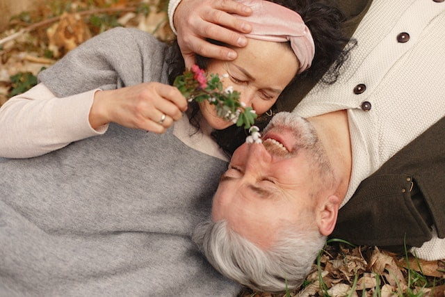 An older couple demonstrate anti-aging tips by laying on the ground with a flower in their mouth.