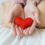 A woman's hands holding a red heart on a bed, promoting wellness tips.