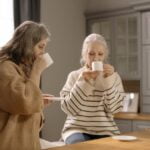 Two older women, engaged in a pleasant conversation, enjoy sipping coffee in the kitchen while sharing anti-aging and wellness tips for senior health.