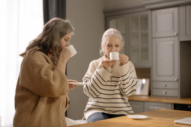 Two older women, engaged in a pleasant conversation, enjoy sipping coffee in the kitchen while sharing anti-aging and wellness tips for senior health.