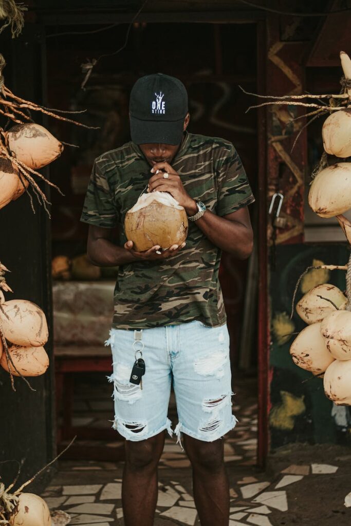 African American male wearing casual outfit drinking fresh coconut water standing near entrance of house