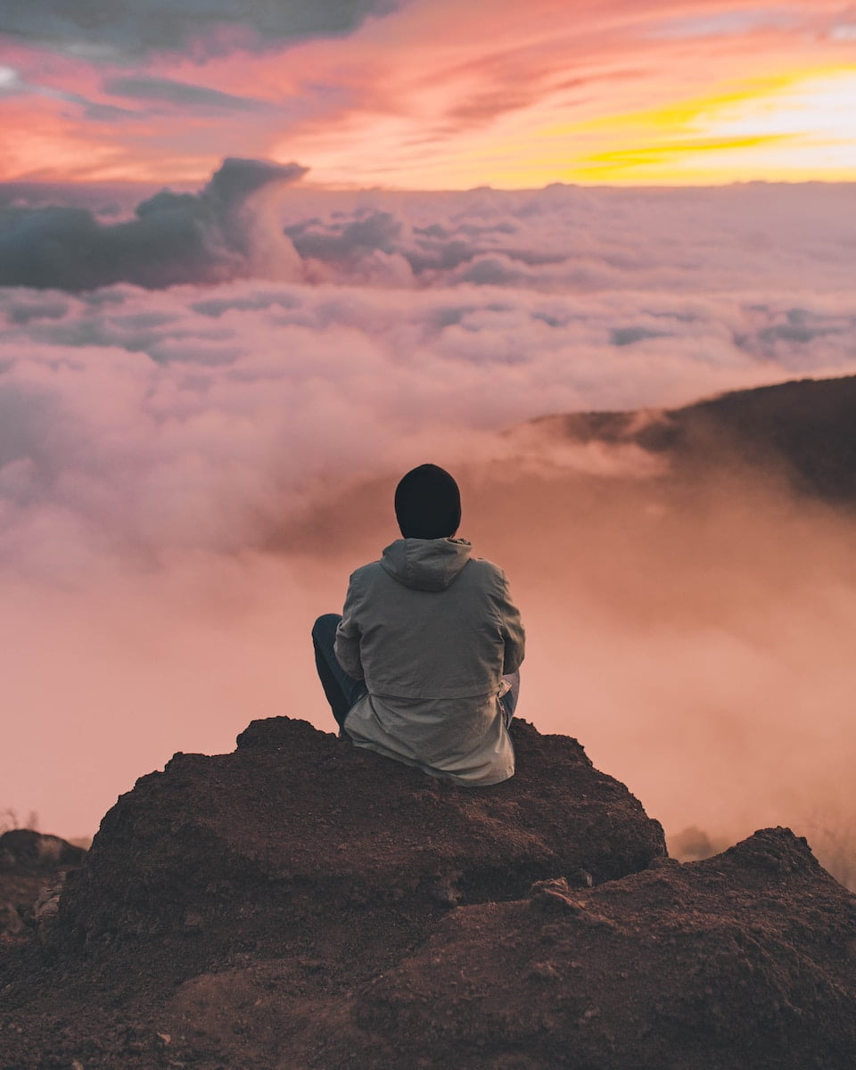A man sitting on top of a mountain overlooking the clouds, finding solace in the serene beauty of nature.