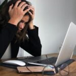 A woman is holding her head in front of a laptop while seeking wellness tips.